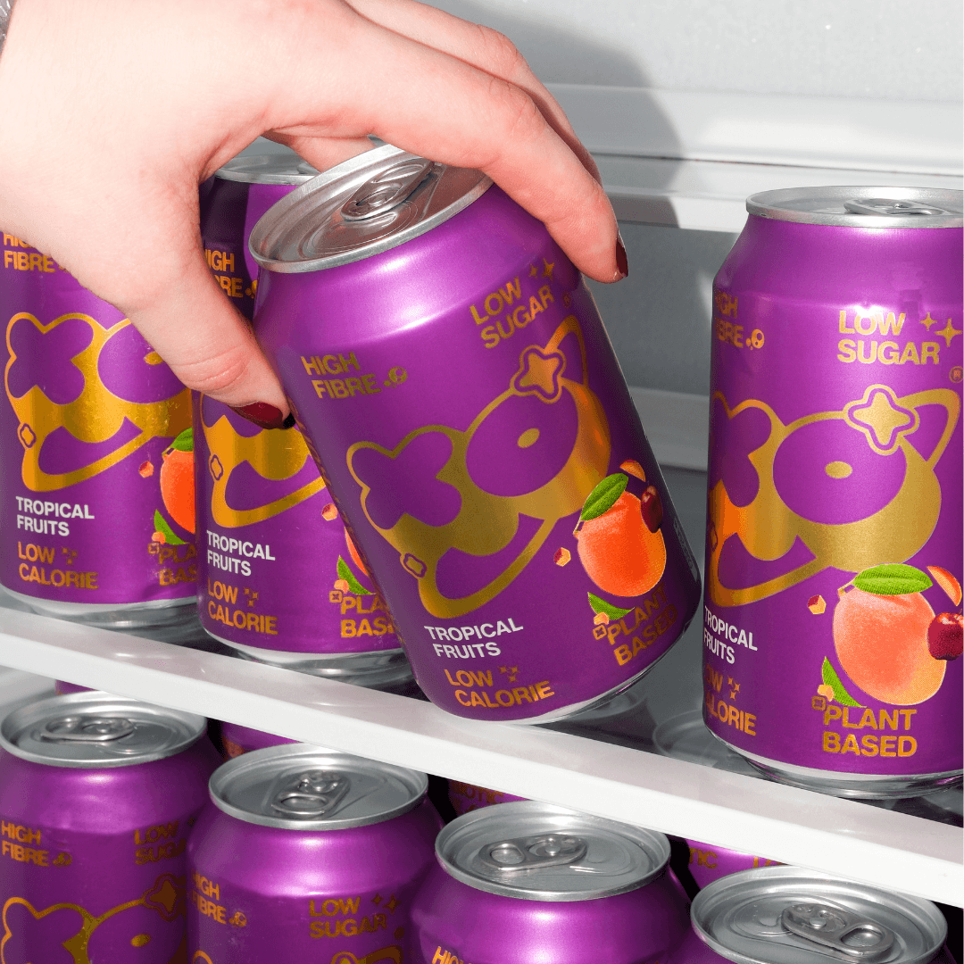 A hand grabbing a can of XO Tropical Soda pack from a colorful fridge shelf.